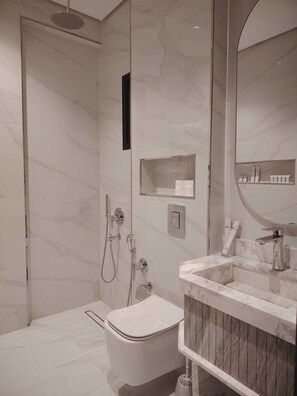 A modern bathroom with a toilet, a shower with a handheld showerhead, a vanity with a mirror, and a shelf with toiletries. - Rama Dubai Serviced Apartments Hafsa (Riyadh)