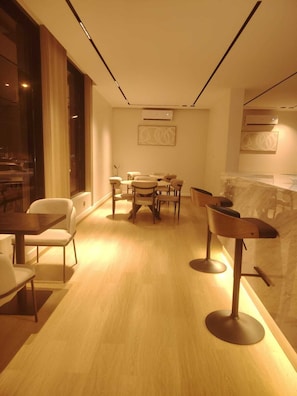 A modern dining area with a wooden table and chairs, a bar area with stools, and a large window with curtains. - Rama Dubai Serviced Apartments Hafsa (Riyadh)