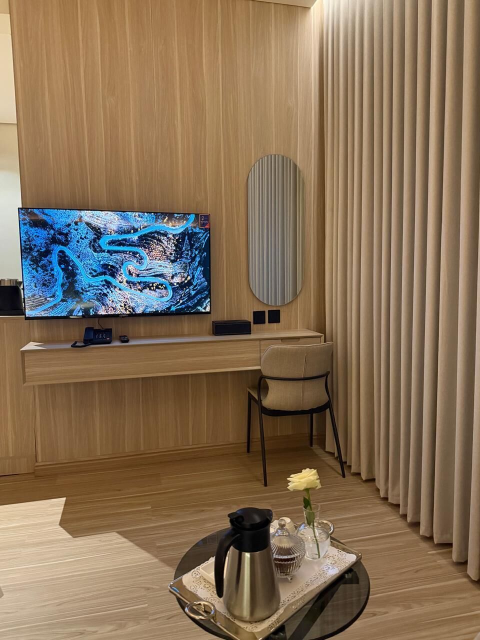 Una camera moderna con una grande TV a schermo piatto, una scrivania in legno, una sedia e un vassoio con una caffettiera e un fiore.