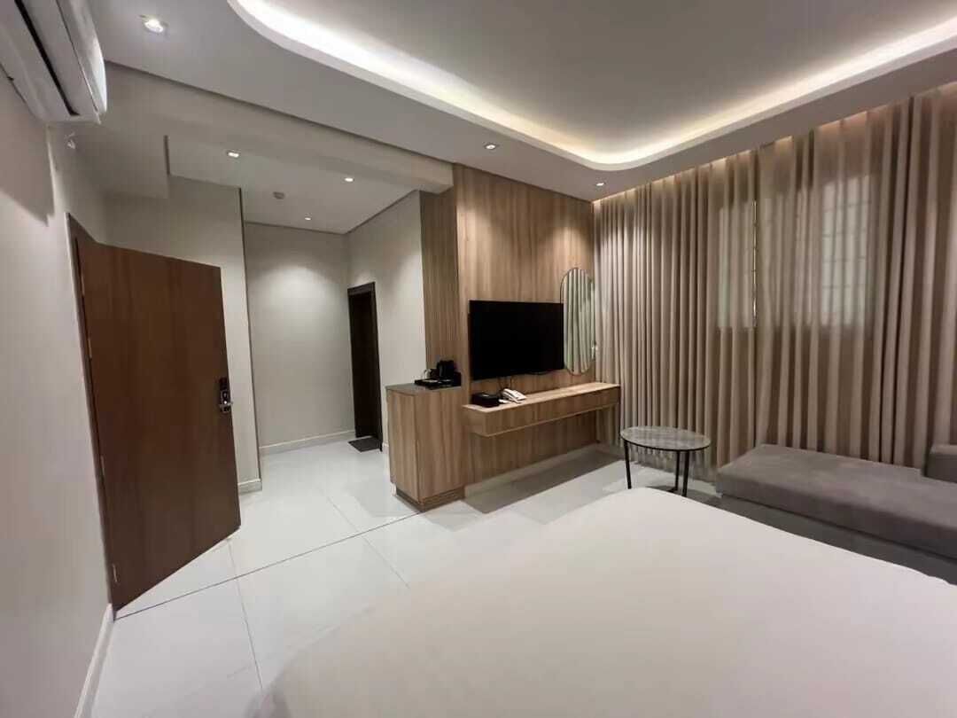 A modern hotel room with a large bed, a TV mounted on a wooden panel, a small round table, and a door leading to another room.