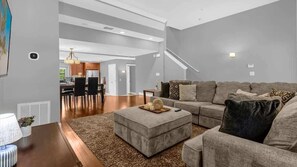 Ein modernes Wohnzimmer mit einem Ecksofa, einem Hocker und einem Essbereich im Hintergrund.