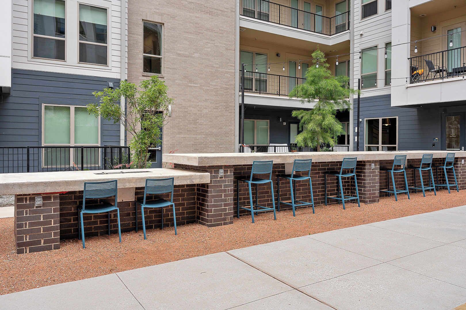A building with a brick exterior, multiple windows, and a row of blue chairs along a concrete ledge.