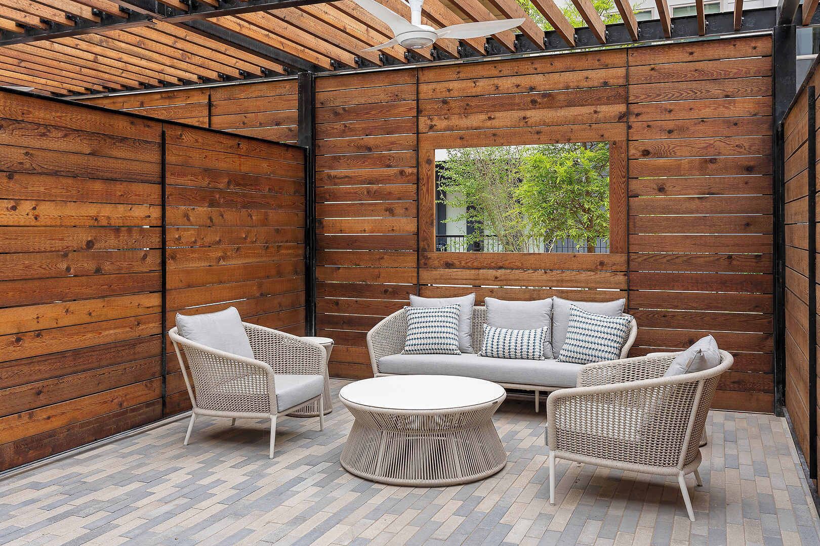Un patio couvert aménagé avec des meubles en osier, une table ronde et une vue sur la verdure par une fenêtre.