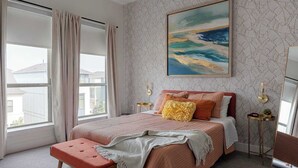 Um quarto com cama grande, um banco, mesa de cabeceira, relógio e janela com cortinas.