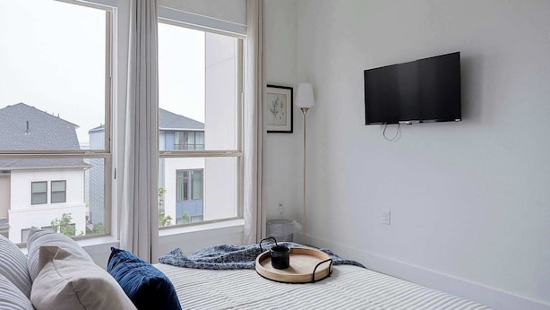 Ett sovrum med en säng, en tv som är monterad på väggen och ett fönster med utsikt över de omkringliggande husen.
