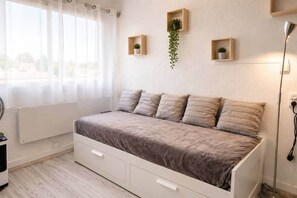 Ein Schlafzimmer mit Bett, Heizkörper, einem fenster mit Vorhängen und Wandregalen mit Pflanzen.