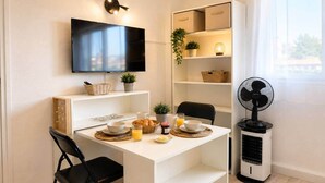 Ein kleiner, moderner Raum mit Fernseher, einem Esstisch mit Frühstücksangeboten, einem Ventilator und Ablageflächen.