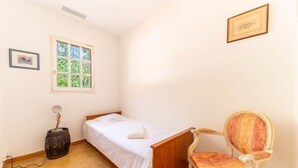 A small, single-bed room with a wooden bed, a chair, a window with a view of greenery, and a wall-mounted picture. - La Villa Peace & Goodness (Vidauban)
