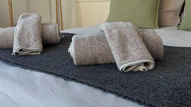 Un lit bien fait, avec des serviettes pliées, un porte-manteau en bois, un coussin vert et un miroir rond accroché au mur.