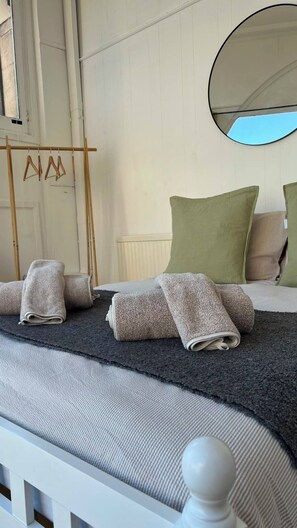 Een netjes opgemaakt bed met opgerolde handdoeken, een houten kledingrek, een groen kussen en een ronde spiegel aan de muur.