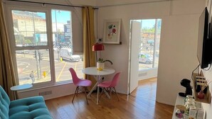 Un salon moderne avec un canapé de couleur turquoise, un coin repas avec des chaises roses, une table blanche et une grande fenêtre donnant sur la rue.