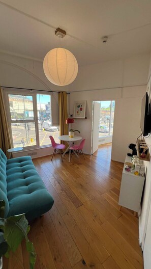 Ein modernes Wohnzimmer mit einem türkisfarbenen Sofa, einem Essbereich mit rosa Stühlen, einem weißen Tisch und einem großen Fenster mit Blick auf die Straße.