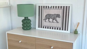 Une commode blanche avec des tiroirs, une lampe verte et, sur le dessus, un tableau encadré représentant un tigre.