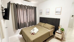 Habitación de hotel con cama, una mesita de noche con planta, televisor montado en la pared y ventana con cortinas.