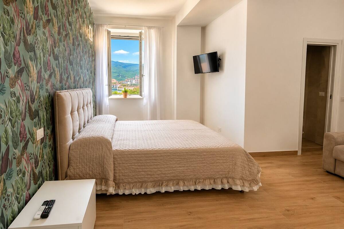 Ein Schlafzimmer mit Bett, einem an der Wand befestigten Fernseher, einem Fenster mit Aussicht und einer Tür.