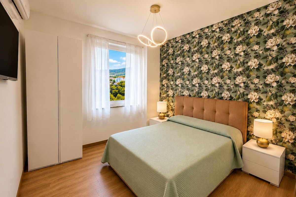 Una camera da letto con un letto, comodini, una finestra con tende, carta da parati a motivi floreali e un lampadario al soffitto.