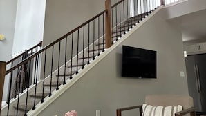 A staircase with a wooden railing, a flat-screen TV mounted on the wall, and a dining area with a table and chairs.