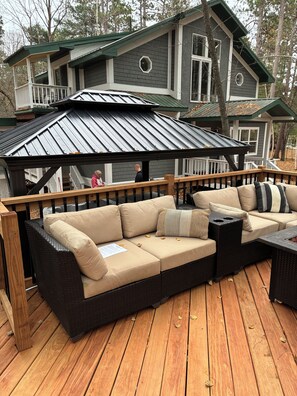 En terrasse med en sofa og et bord, omgivet af træterrasse og et hus i baggrunden.