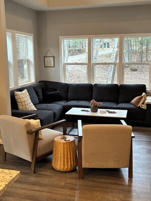 A living room with a sectional sofa, a coffee table, and two armchairs. There are large windows with a view of trees and a path. - HOT TUB! New 2027  - Newly Remodeled Modern house with AC in Crosslake (Crosslake)
