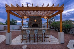 An outdoor barbecue area with a stone fireplace, wooden pergola, and seating. - Havasu Acoma - Pool, Spa, Game Room, Sleeps 16 (Lake Havasu City)