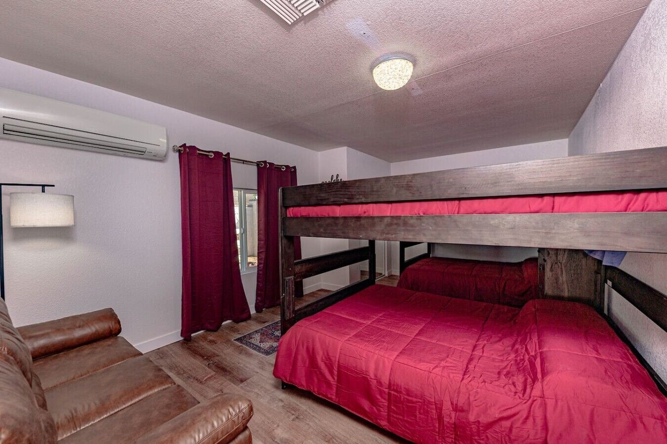 Un letto a castello con biancheria rossa, un divano in pelle marrone, una lampada e una finestra con tende.