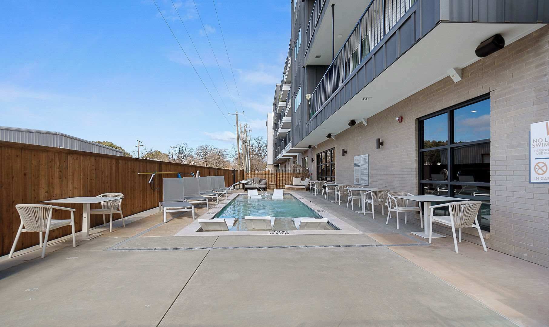 A pool area with lounge chairs and tables, a wooden fence, and a building with a sign indicating no diving.