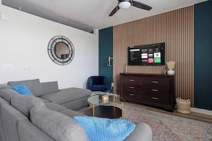 A modern living room with a grey sofa, a blue armchair, a glass coffee table, a dark wooden TV stand, and a wall-mounted TV displaying various streaming services. - DallasHaus | Pool & Gym, Love Field (Dallas)