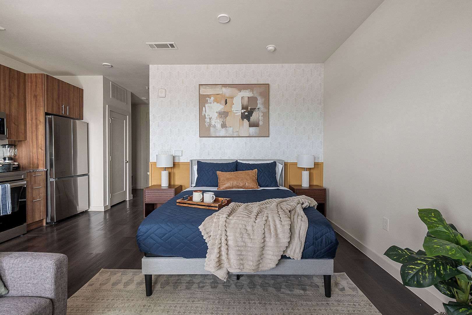 A modern bedroom with a bed, bedside tables, a painting, and a kitchen area.