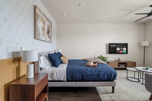 Ein modernes Hotelzimmer mit Bett, Nachttisch, Lampe, Fernseher und einem kleinen Beistelltisch mit Pflanze.