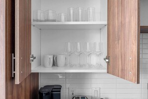 A kitchen with wooden cabinets, a white countertop, and a shelf with clear drinking glasses and mugs. - DallasHaus | Pool & Gym, Love Field (Dallas)