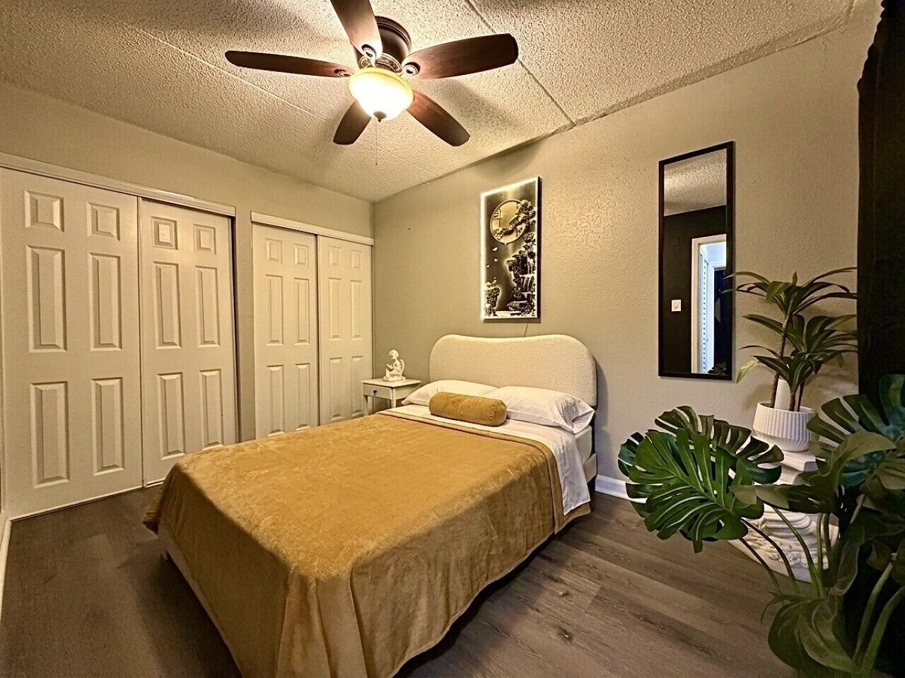 A bedroom with a bed, a ceiling fan, a nightstand, a mirror, and a potted plant.