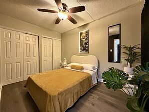 A bedroom with a bed, a ceiling fan, a nightstand, a mirror, and a potted plant.