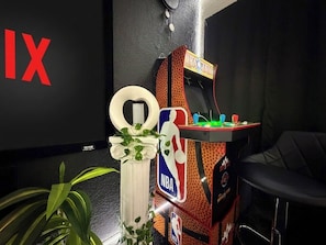 Una habitación con una máquina recreativa, un jarrón blanco con una planta y una silla negra.