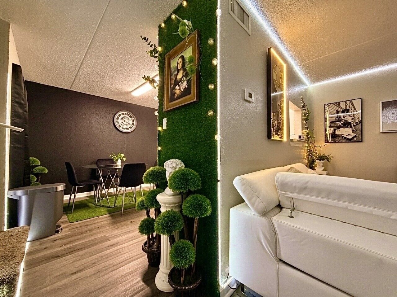 A modern living room with a white sofa, a dining area with a round table and chairs, and a green wall with a framed picture.