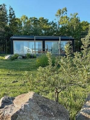 Uma casa moderna de vidro com telhado plano, cercada por uma área gramada com pedras e arbustos.