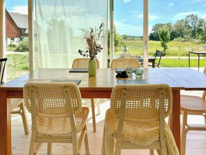 Uma área de refeições com mesa de madeira e cadeiras de vime, um vaso com flores, e vista para uma paisagem verdejante através de grandes janelas.