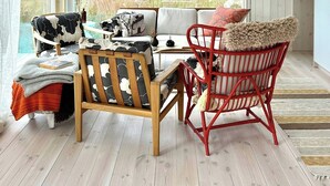 Een kamer met een houten plafond, een groot raam met uitzicht op groen, een bank met decoratieve kussens, een houten stoel en een rode stoel met een zachte kussen.