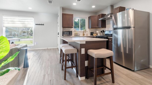 A modern kitchen with a stainless steel refrigerator, wooden cabinets, a bar stool seating area, and a window with blinds. - East Austin Retreat w/ Hot Tub & Backyard (Austin)