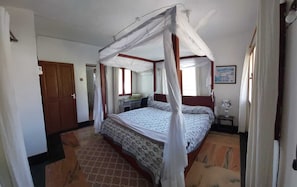Une chambre à coucher avec un lit à baldaquin, un bureau avec une chaise, une petite table avec une lampe et une porte donnant sur une autre pièce.