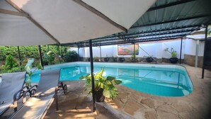 Una piscina con sillas de descanso y plantas en macetas, bajo una zona cubierta con techo de metal.