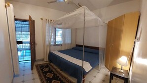 Un dormitorio con una cama, un ventilador de techo, una mosquitera, una mesita con una lámpara y vista a una piscina a través de una ventana.