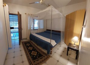 Et soveværelse med en seng, en loftsvift, et myggenet, et lille bord med en lampe og udsigt til en pool gennem et vindue.
