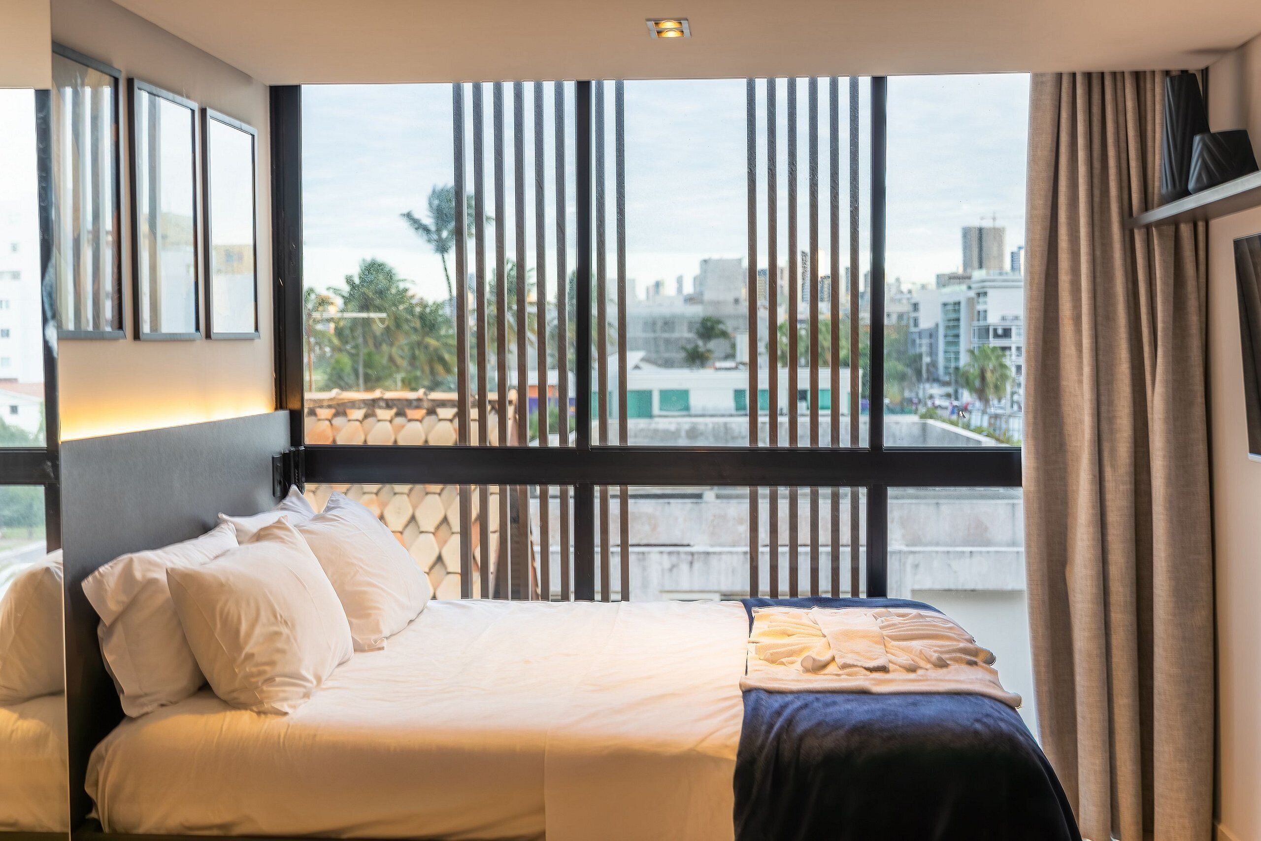 A hotel room with a large window, a bed with white linens, and a view of the cityscape.