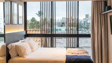 A hotel room with a large window, a bed with white linens, and a view of the cityscape.