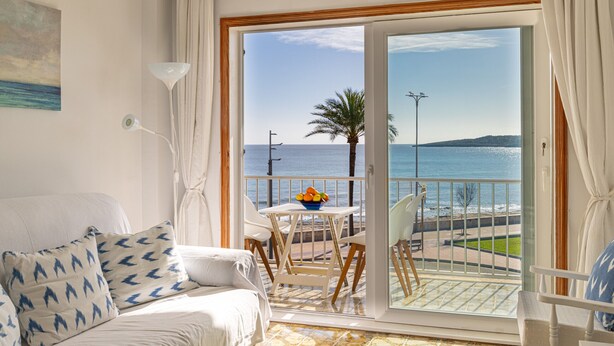 En balkon med udsigt over havet, et spisebord med stole og en sofa med mønstrede puder.