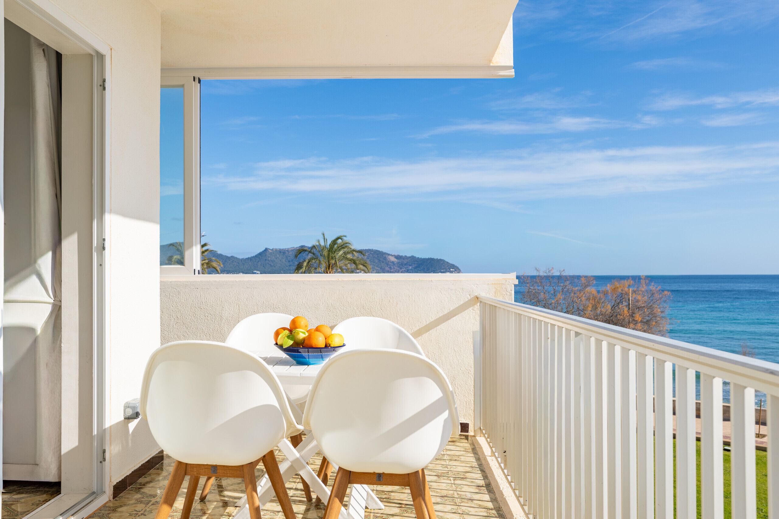Un balcón con mesa y sillas, un frutero y vistas al mar y a las montañas.