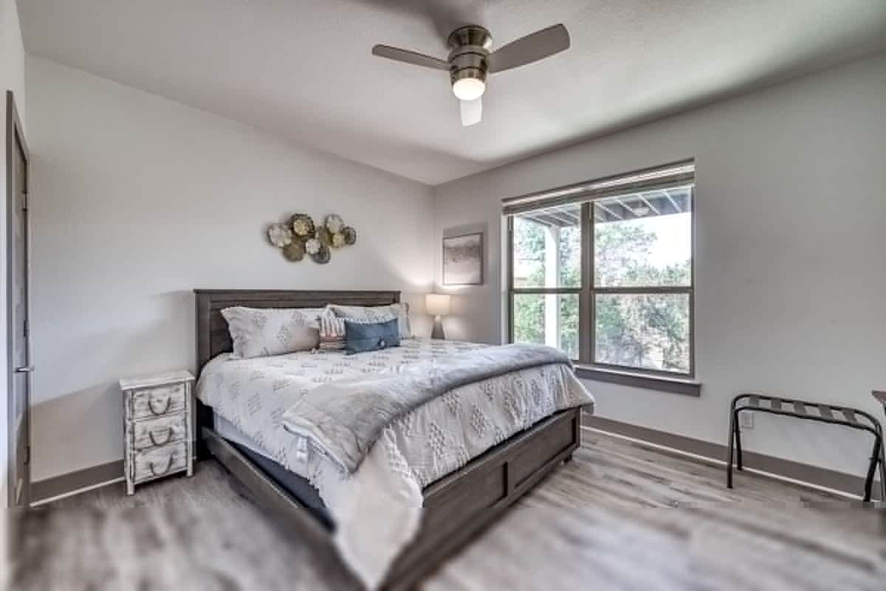 A bedroom with a bed, a nightstand, a ceiling fan, a window, and a wall-mounted decoration.