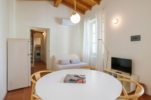 Ein Zimmer mit weißem Tisch, Stühlen, einer Couch, einem Fernseher und einer Klimaanlage.