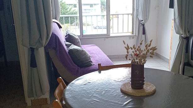 Une pièce avec une table ronde, des chaises, un canapé violet, une fenêtre avec des rideaux et une vue sur l’extérieur.