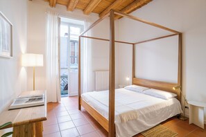 Ein Bett mit Baldachin, weiße Bettwäsche, eine Nachttischlampe und Blick auf einen Balkon mit Geländer.